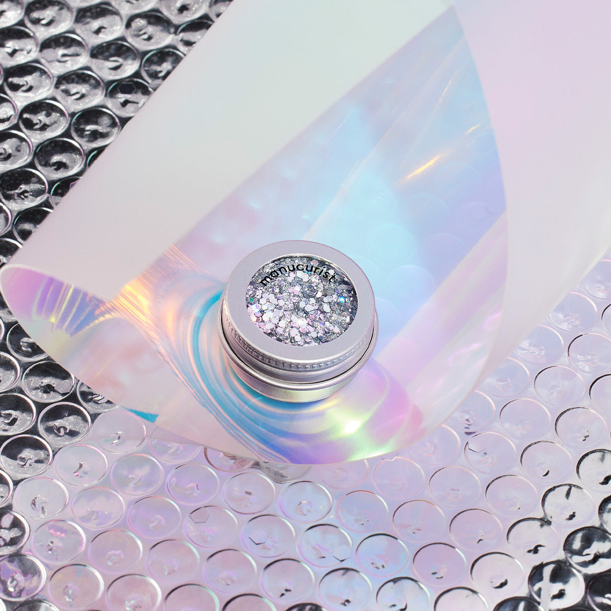 Un petit pot de paillettes biodégradables Holo Manucurist posé sur du papier bulle irisé, partiellement recouvert d&