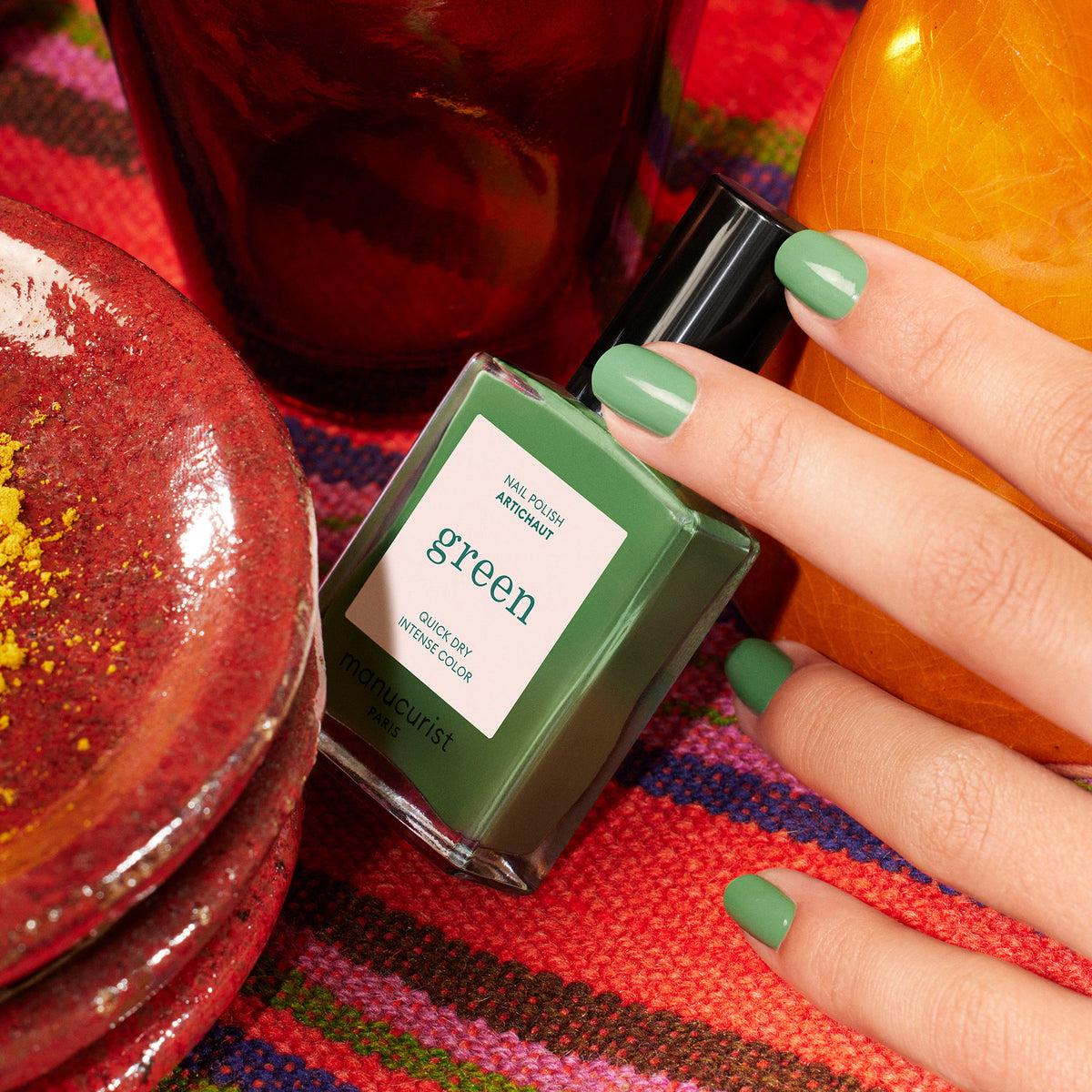 A hand with green-painted nails holds a Manucurist nail polish bottle labeled Artichoke, resting on vibrant woven fabric and surrounded by red and orange ceramics for a Moroccan garden vibe.