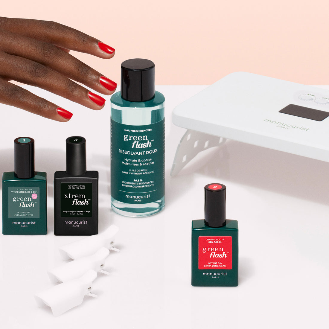 A hand with red-painted nails reaches toward the Manucurist Discovery Kit, which includes Green Flash gel polish, vitamin-rich base coat, remover, nail clips, and a UV lamp, all arranged on a white surface.