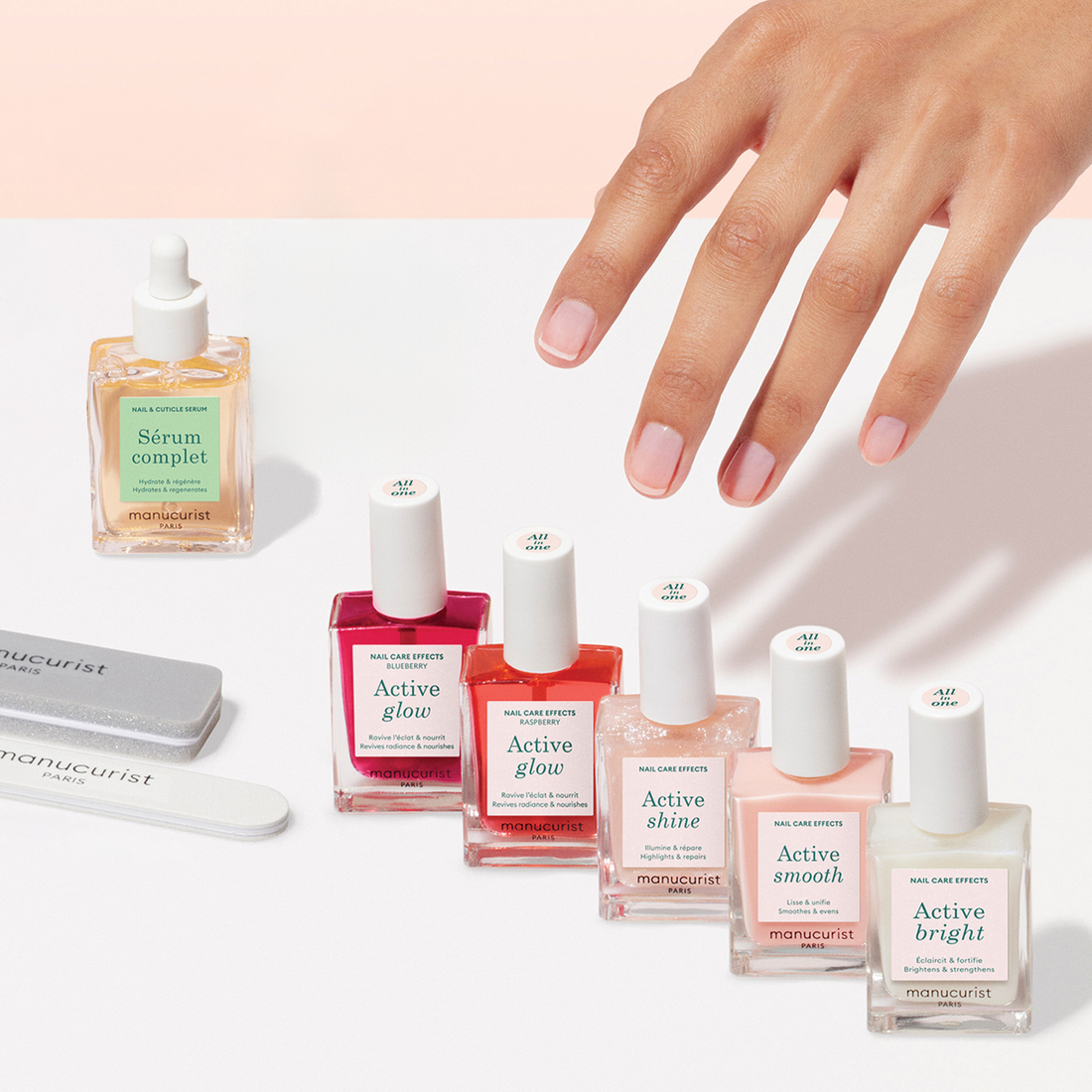 A manicured hand reaches for the Super Active Manicure Kit by Manucurist US, featuring vibrant polishes, nourishing serums, and nail files—the ultimate manicure set—arranged on a pale pink background.