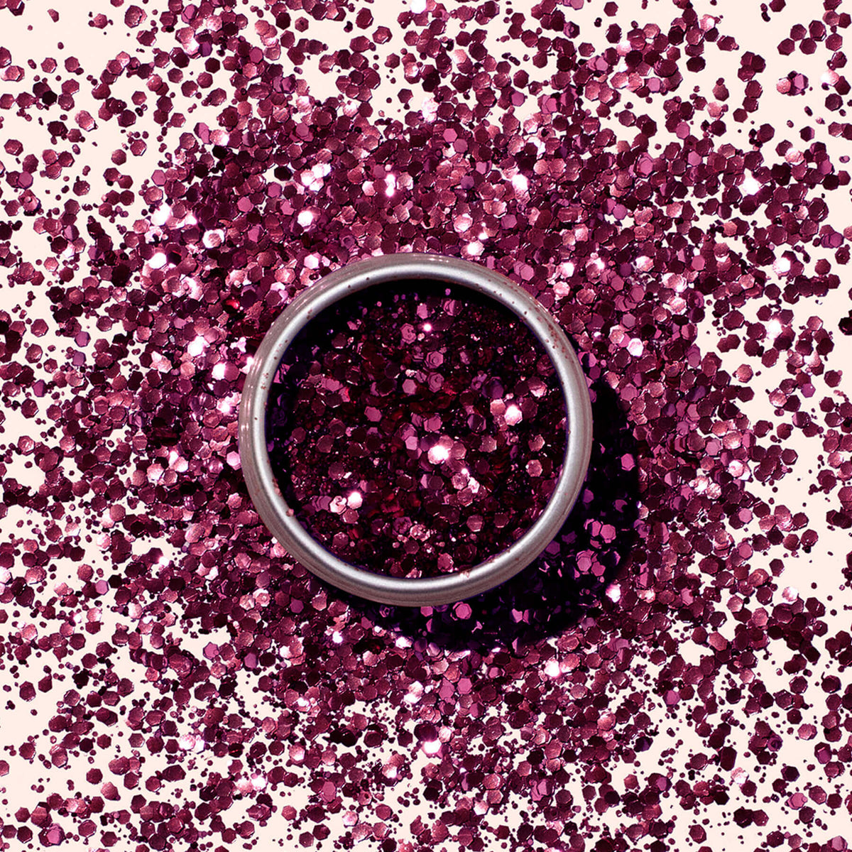 Une boîte ronde en métal de Paillettes biodégradables Bordeaux de Manucurist est posée sur une surface, entourée de paillettes magenta dispersées qui scintillent selon un motif circulaire assorti au produit écoresponsable.