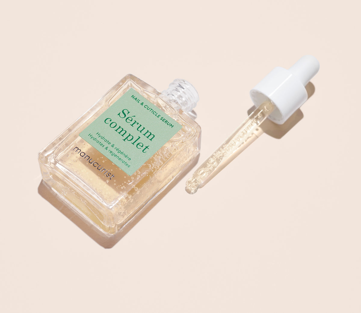 A clear glass bottle of Serum Complet nail and cuticle serum with a pale green label lies next to its dropper cap, both on a light beige background. The dropper contains a small amount of the serum.