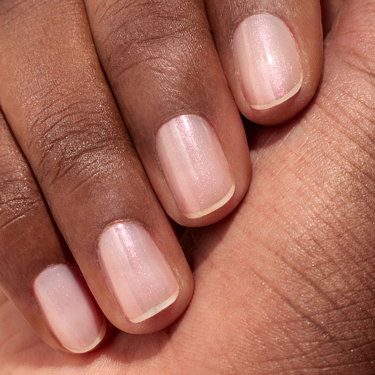 Gros plan sur une main à la peau mate, dévoilant des ongles impeccablement manucurés avec le Glitters Duo Rose Gold Glazed de Manucurist — un vernis rose pâle translucide au délicat reflet nacré.