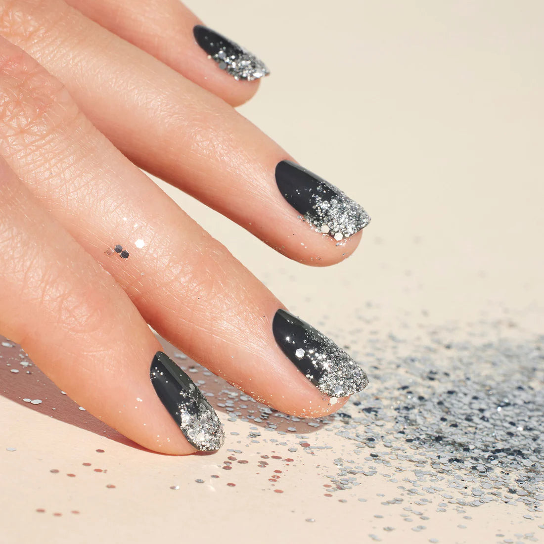 Une main avec un vernis à ongles noir aux pointes ombrées pailletées argentées repose sur une surface claire, entourée de paillettes argentées éparpillées.