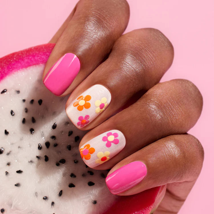 A hand with pink and white nails, some decorated with colorful flower designs, holds a slice of dragon fruit against a pink background.