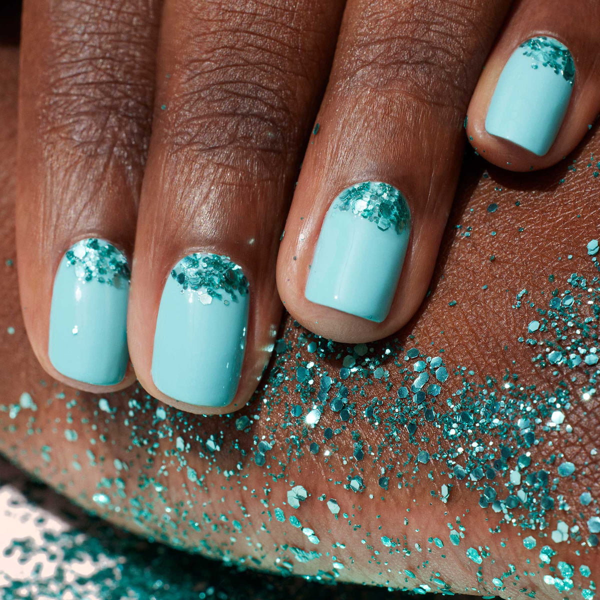 Gros plan sur une main au vernis bleu clair, ornée de Paillettes Biodégradables Manucurist turquoise près des cuticules appliquées avec un pinceau Nail Art. Quelques paillettes sont également parsemées sur la peau sous la main.