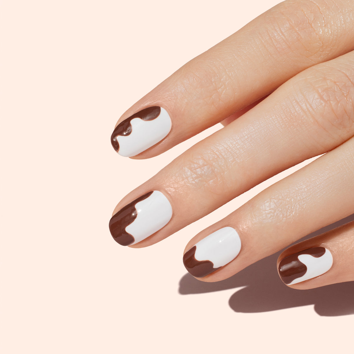 A hand with light skin tone showing four fingers, each with white nail polish and brown, wavy drip designs near the tips, against a pale peach background.