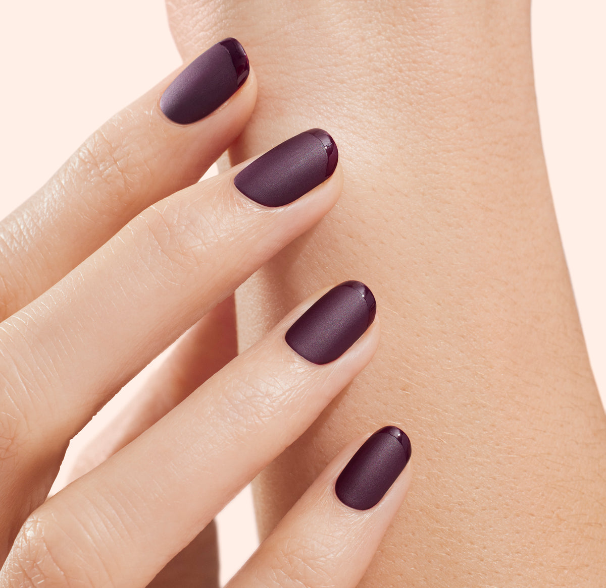 A close-up of a hand with neatly manicured nails painted in a matte dark purple shade, lightly touching another part of the hand against a light background.