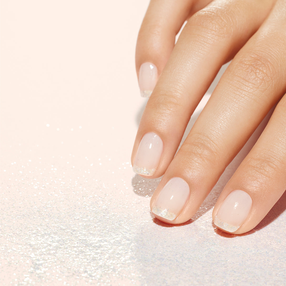 A close-up of a hand with natural, short nails featuring a sheer pink polish and glittery tips, resting on a light pink surface scattered with fine, sparkling glitter.