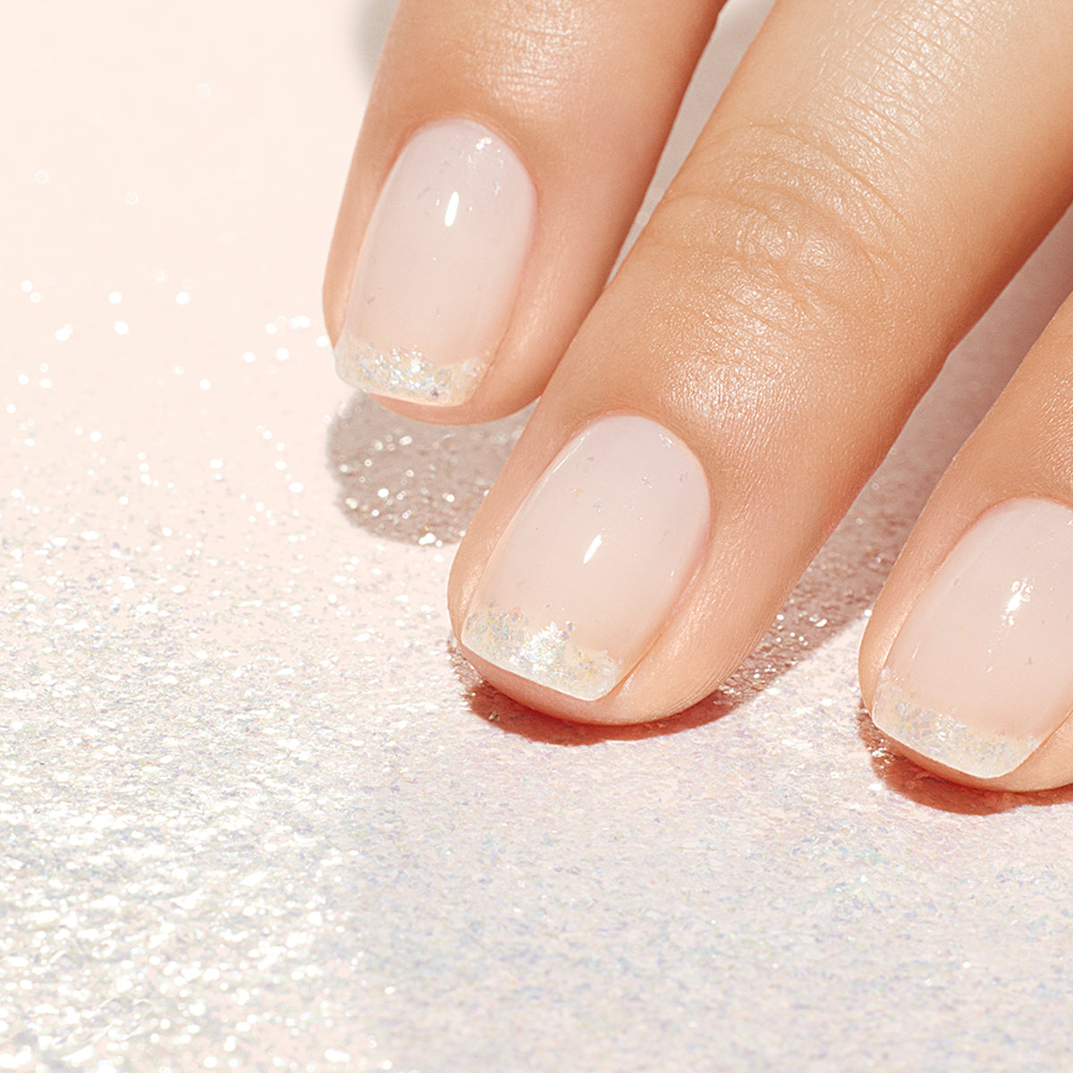 A close-up of a hand with a Manucurist Pearlescent Glitter Flakes manicure; short nails in pale pink, adorned with delicate silver and pearlescent flakes at the tips. A sparkly background enhances the elegant look.