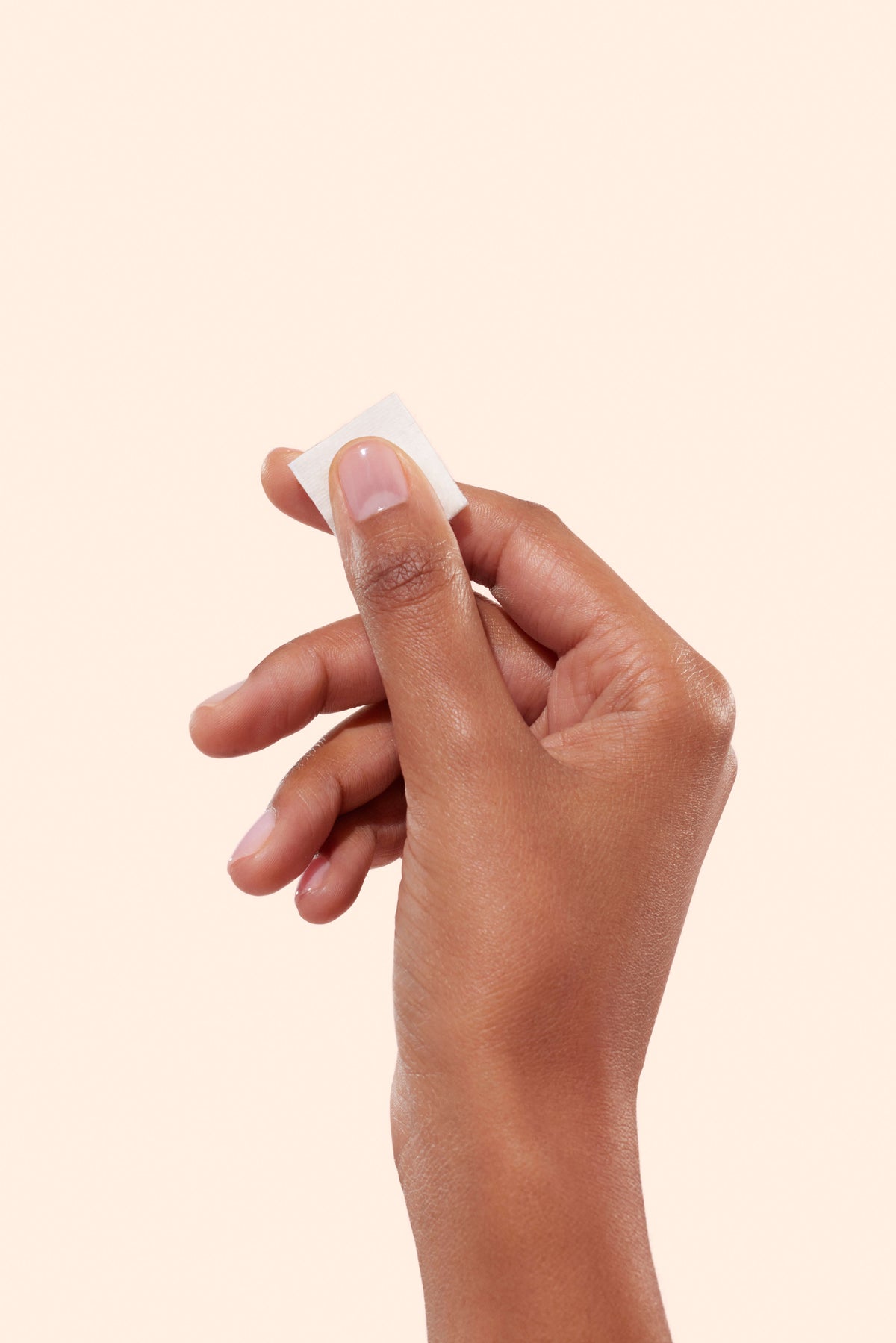A hand with medium brown skin holds a small white square cotton pad between the thumb and index finger against a plain light beige background.