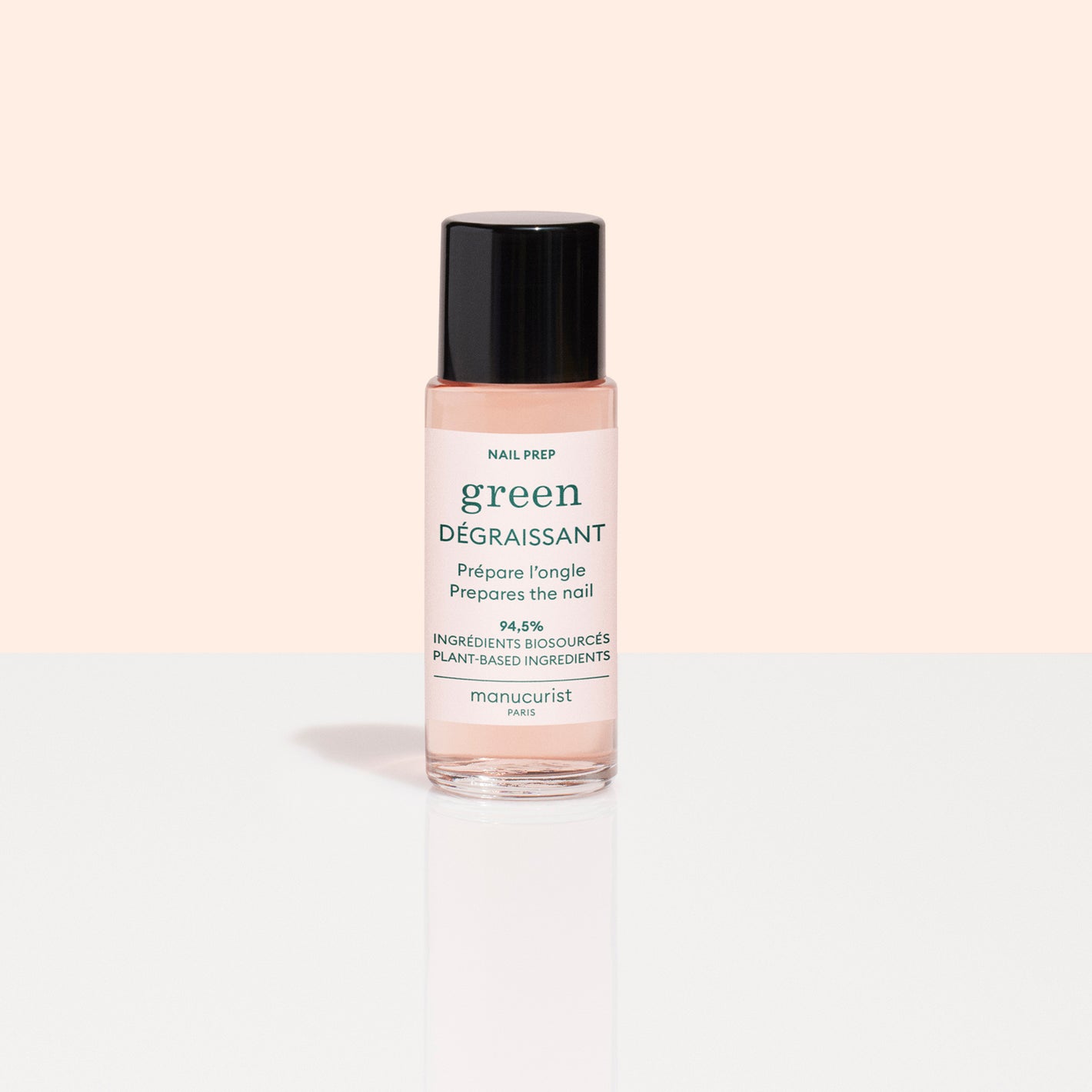 A small bottle of Manucurist Degreaser, a plant-based nail prep with clear pink liquid and black cap, sits on a reflective white surface. Its white label with green and pink text stands out against a pale peach background.