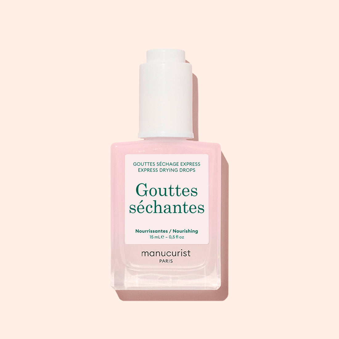 A square bottle of Manucurist Drying Drops with a white cap, featuring light pink liquid, is shown against a pale pink background. This nail product speeds up drying time and nourishes cuticles.