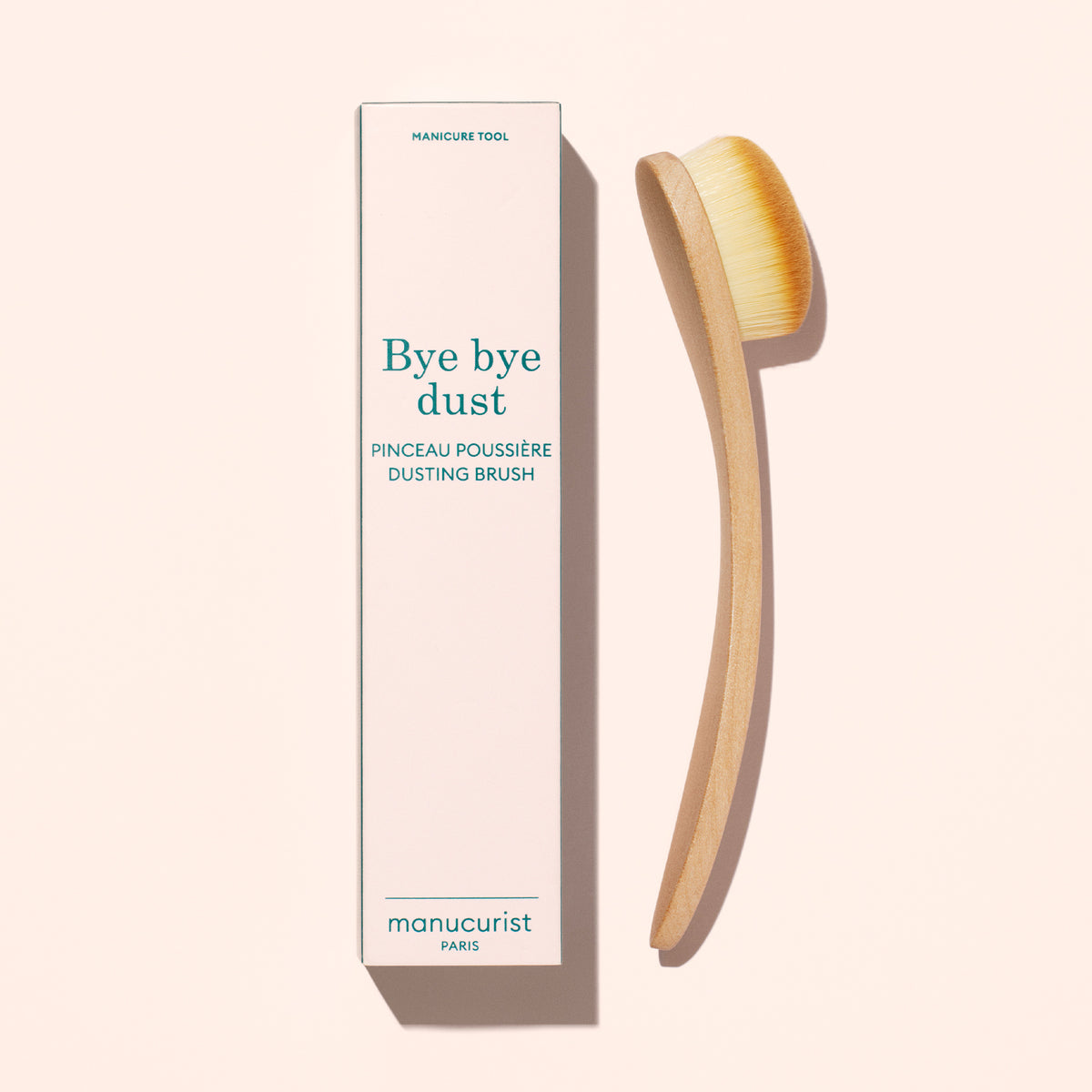 Un pinceau dépoussiérant Dust Brush de Manucurist aux poils vegan et au manche en bois courbé est posé à côté de sa boîte haute portant la mention « Bye bye dust – Dust Brush » sur un fond beige clair.