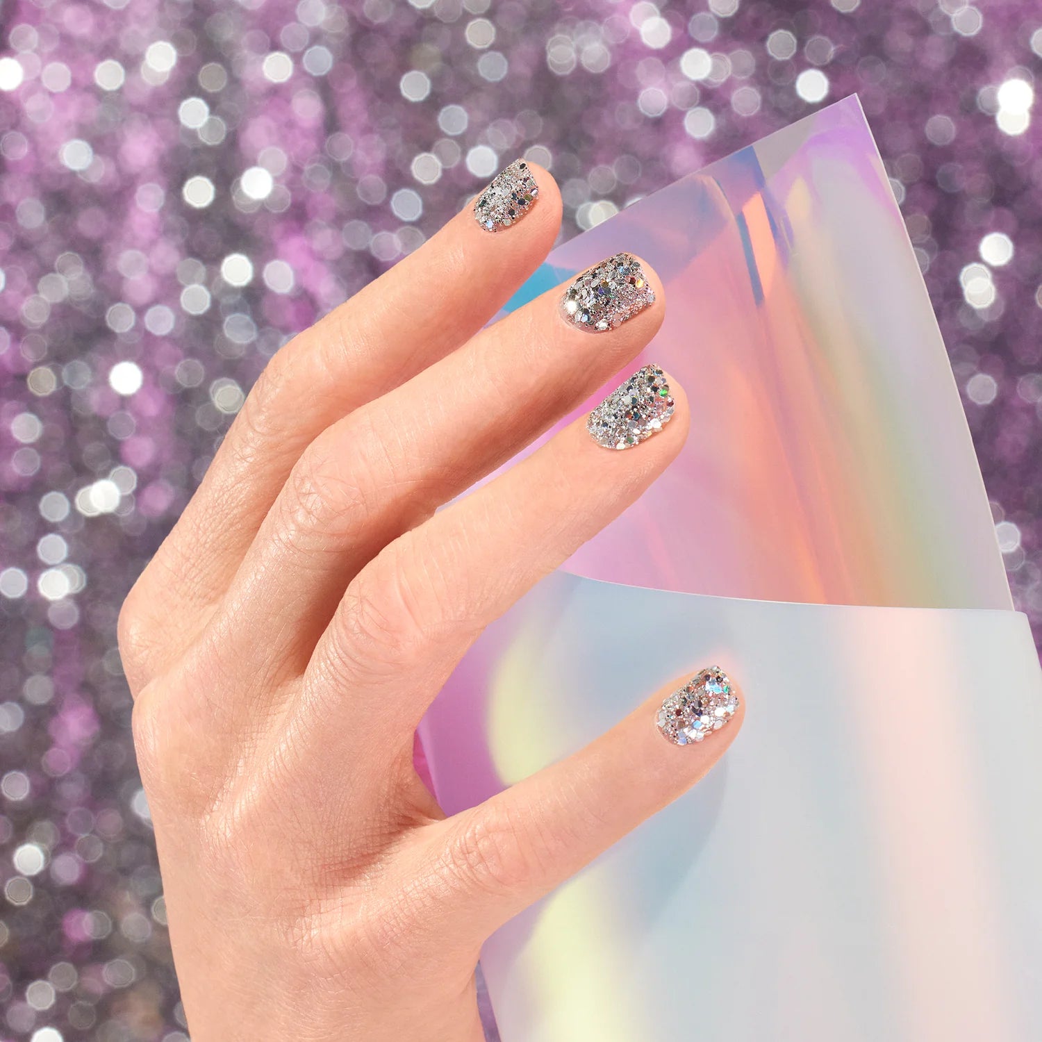 A hand with silver glitter nail polish holds a sheet of iridescent, holographic paper against a sparkly purple background.