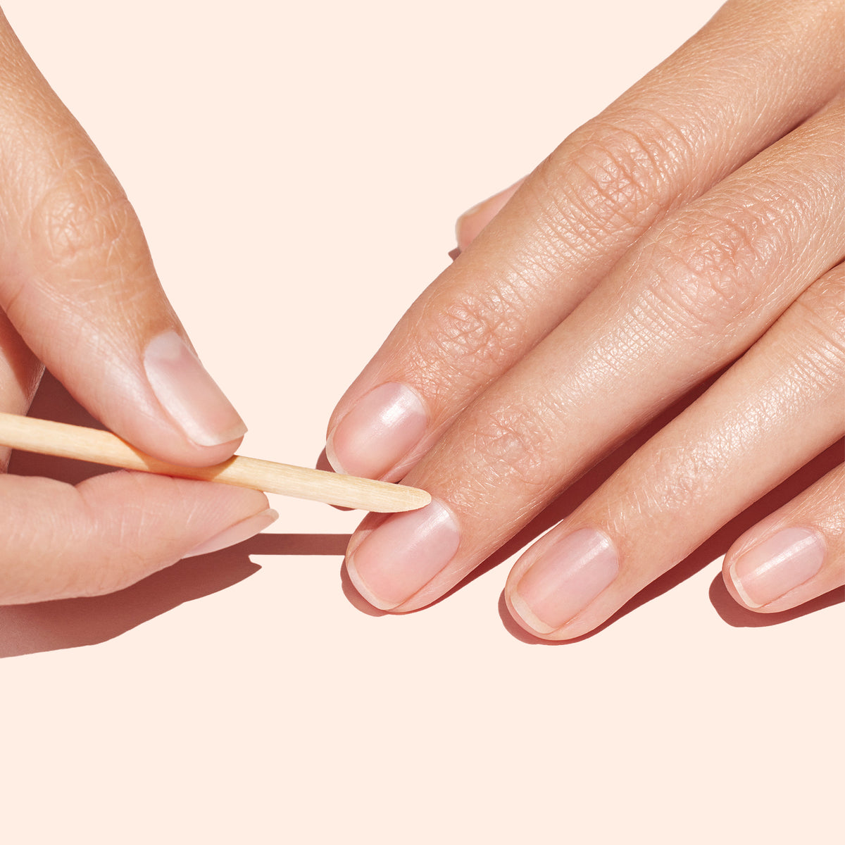 Une main utilise un Bâtonnet repousse-cuticules en bois x20 Manucurist pour repousser délicatement les cuticules d’une autre main sur fond pêche clair, lors d&