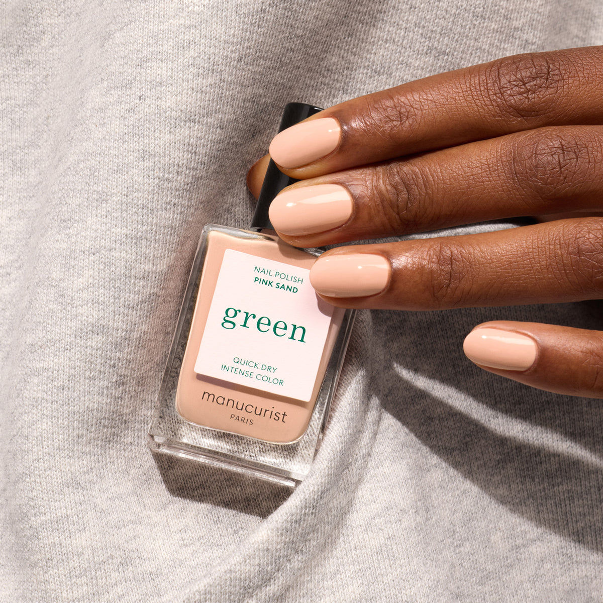 Une main aux ongles parfaitement manucurés, vernis dans la teinte Pink Sand, un nude pâle de Manucurist, tient un flacon de vernis à ongles assorti sur un tissu gris doux.