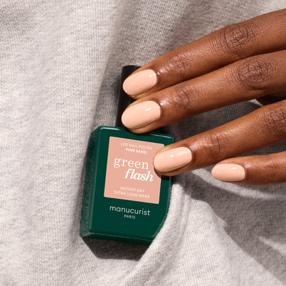 A hand with neatly manicured, light pink nails holds a green Manucurist bottle of Pink Sand gel polish—a soft shade with subtle orange undertones—resting on gray fabric.