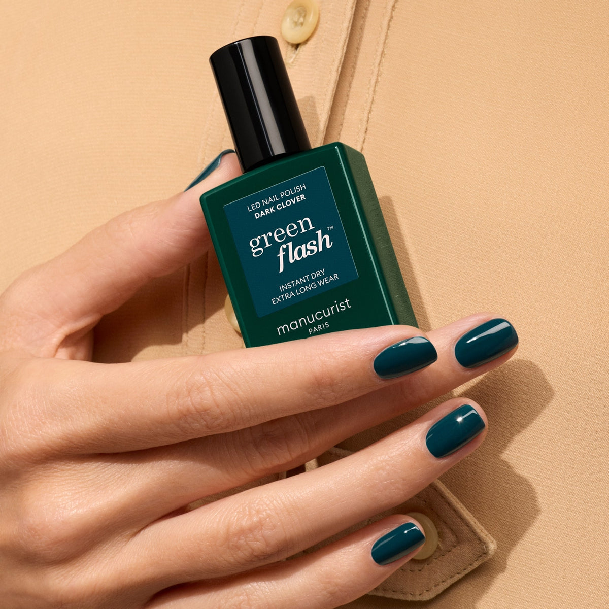 Une main aux ongles courts et soignés vernis en vert foncé tient un flacon de vernis gel Dark Clover de Manucurist assorti, sur fond de chemise beige.