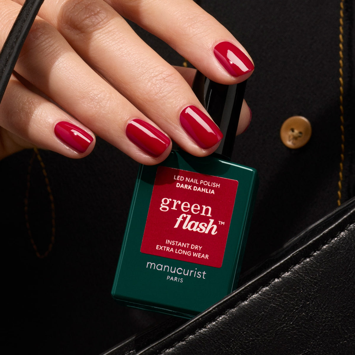 Une main aux ongles rouges brillants tient un flacon de vernis gel Manucurist Dark Dahlia près d&