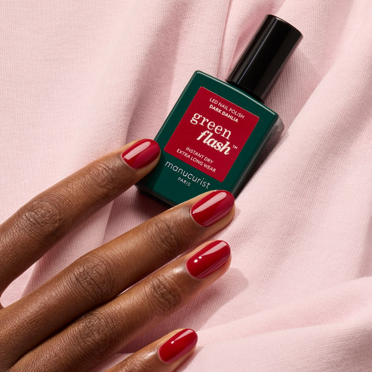 Une main aux ongles rouges soigneusement manucurés tient un flacon de vernis gel Manucurist Dark Dahlia sur un fond de tissu rose poudré.