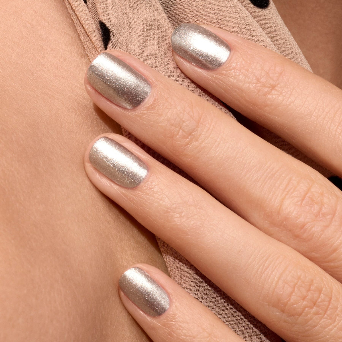 Gros plan sur une main aux ongles courts et soigneusement manucurés, peints avec Disco de Manucurist, un vernis argenté pailleté opaque, reposant sur un tissu beige à pois noirs.
