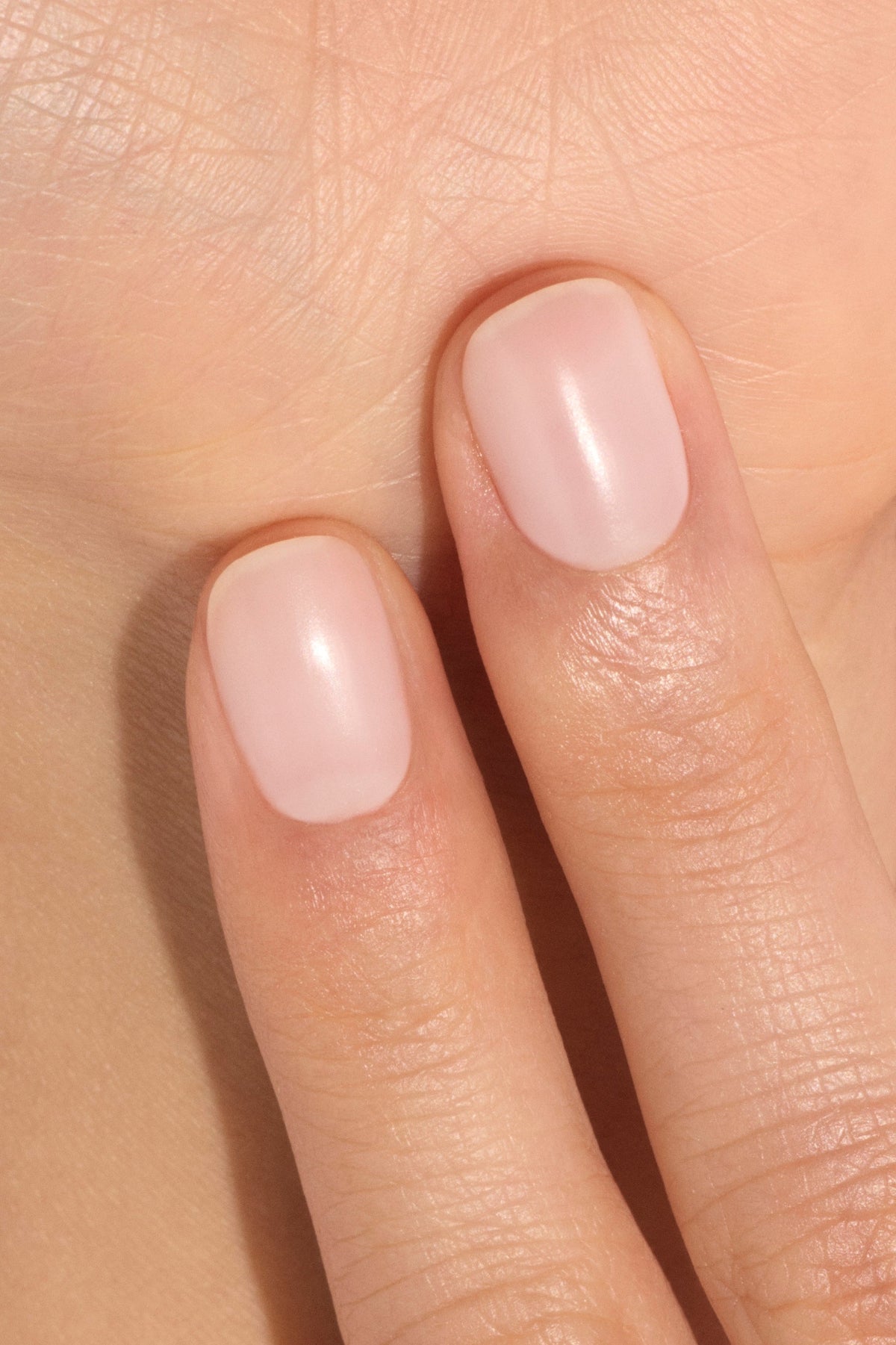 Deux doigts aux ongles soigneusement limés sublimés d'un vernis rose clair naturel touchent la paume d'une main, mettant en valeur des ongles nets et impeccables.