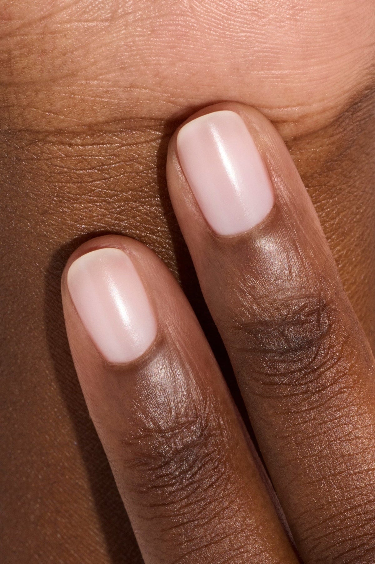 Deux doigts aux ongles soigneusement coupés, vernis d’une teinte rose pâle ou nude, reposent sur une peau brun foncé. Les ongles ont un fini lisse et brillant, et la peau paraît saine et hydratée.
