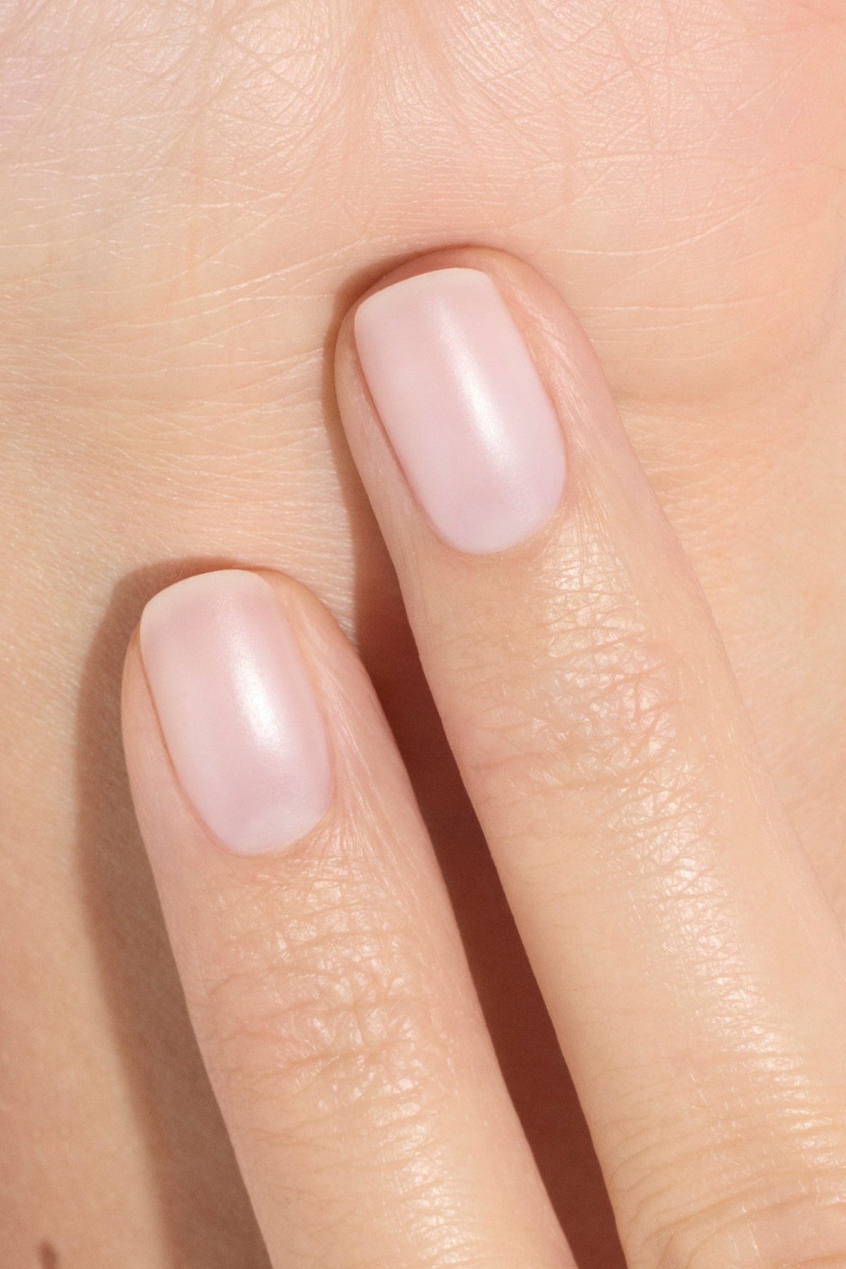 Gros plan sur deux doigts aux ongles courts et impeccablement manucurés, vernis d'une teinte rose clair naturelle, posés contre la paume d'une main. La peau est lisse et bien hydratée.