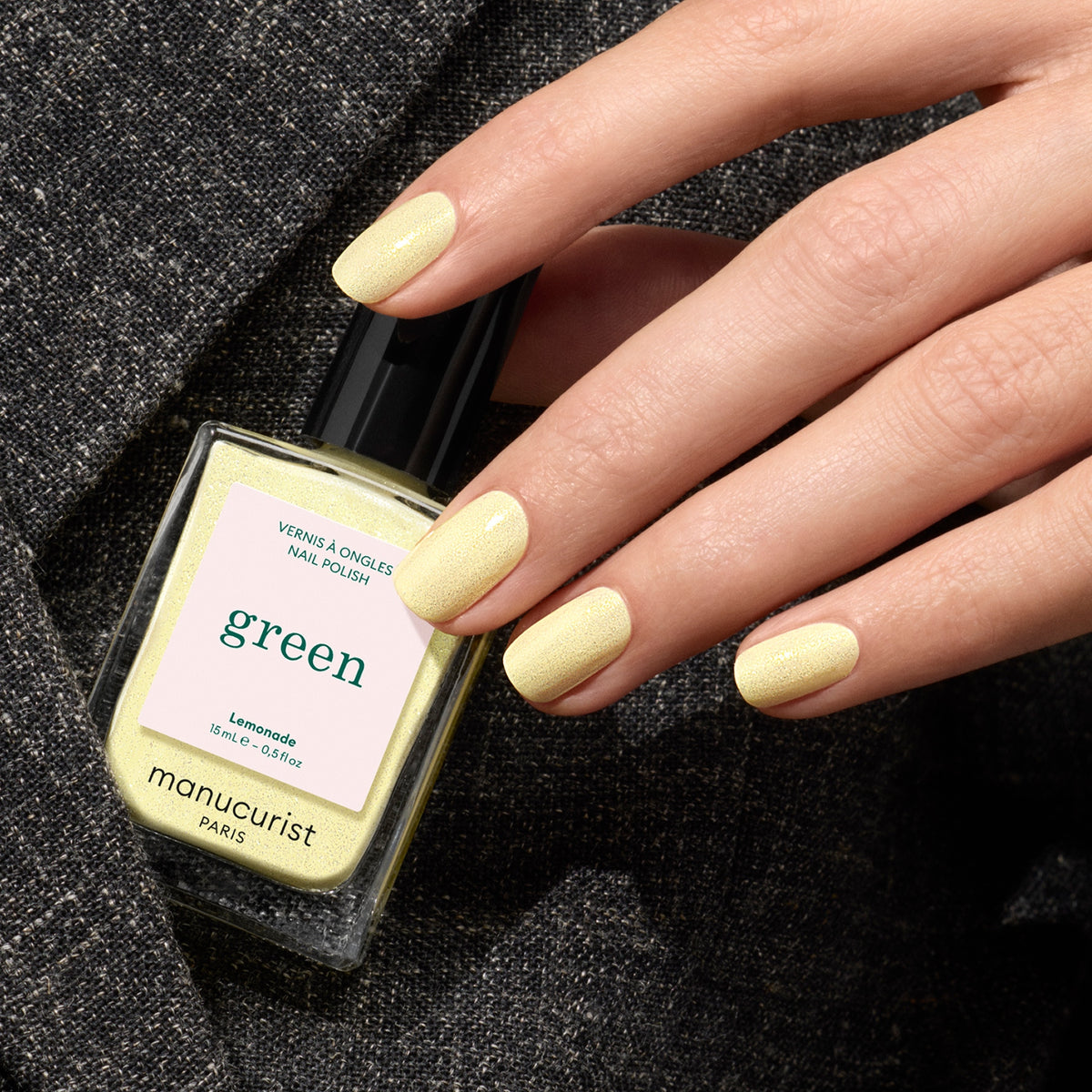 Une main aux ongles soigneusement manucurés et peints en jaune pâle tient un flacon assorti de Lemonade de Manucurist, sur un fond de tissu sombre texturé.