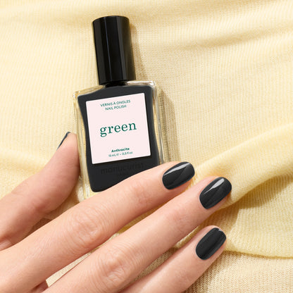 A hand with neatly manicured, glossy dark grey nails holds a bottle of Manucurist Anthracite nail polish against a soft yellow fabric background.
