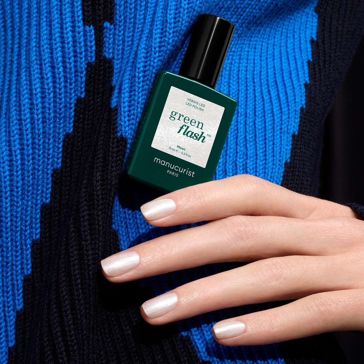 Une main aux ongles blancs nacrés et scintillants tient un flacon de vernis Manucurist Moon sur un fond bleu et noir à motifs.