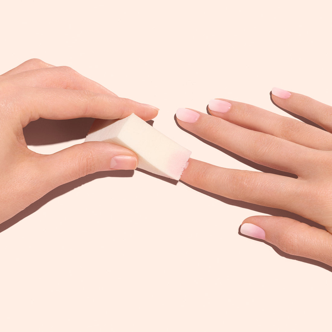 A hand uses Manucurist Nail Art Sponges (x4) to blend light pink polish, creating soft Baby Boomer nails on the other hand against a pale background.