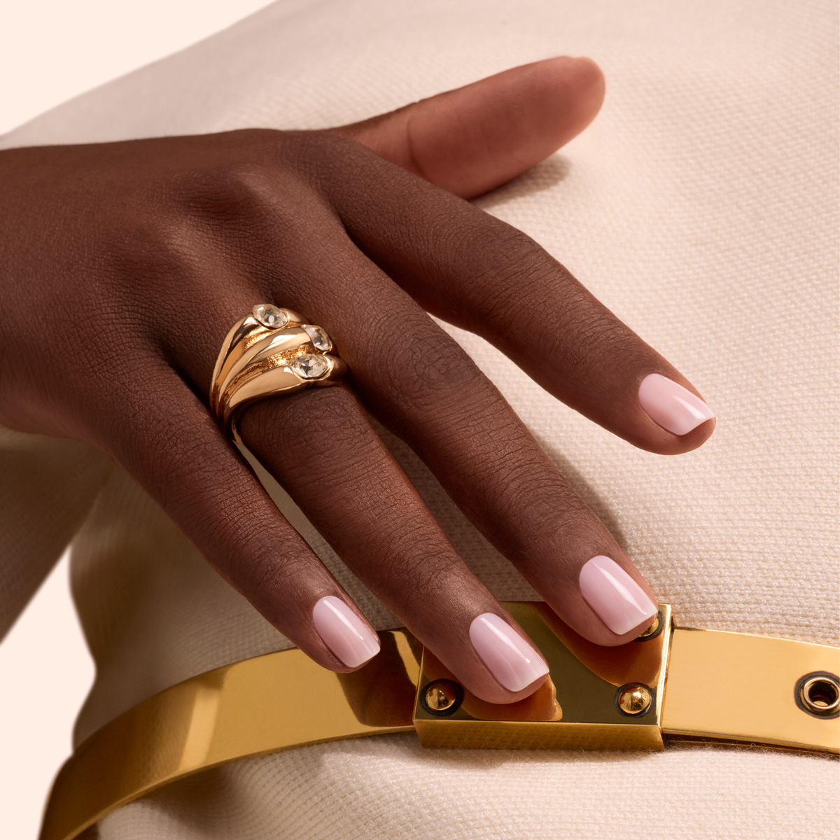 Une main aux ongles vernis avec la teinte Milky Pink de Manucurist repose sur un tissu écru texturé. Elle porte une grande bague en or sertie de trois pierres précieuses. Une ceinture dorée brillante est également visible.