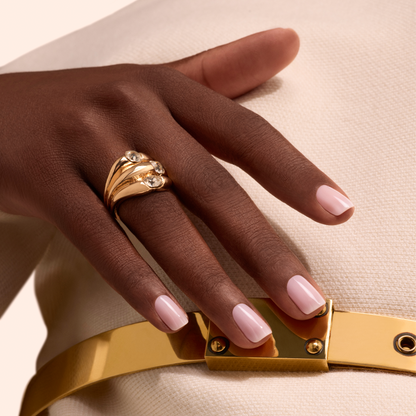 Une main aux ongles vernis avec la teinte Milky Pink de Manucurist repose sur un tissu écru texturé. Elle porte une grande bague en or sertie de trois pierres précieuses. Une ceinture dorée brillante est également visible.