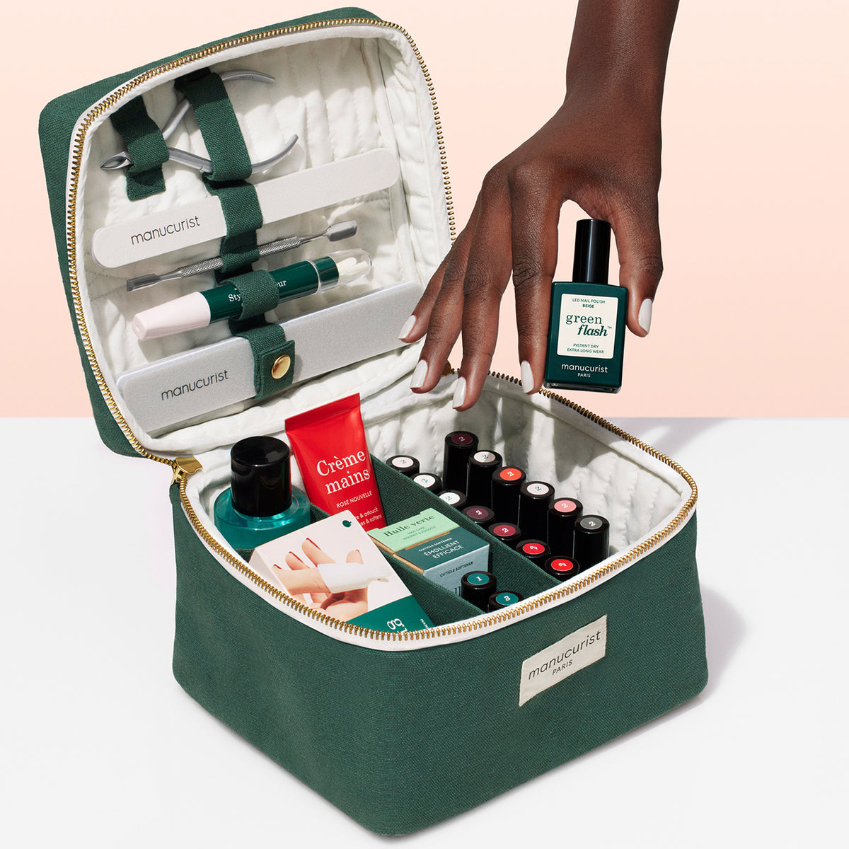 La Vanity Case Manucurist-test est un kit de manucure vert avec des outils de soin des ongles, des vernis, une crème pour les mains et des accessoires dans une pochette zippée — un organisateur compact. Une main tient un vernis « green flash » .
