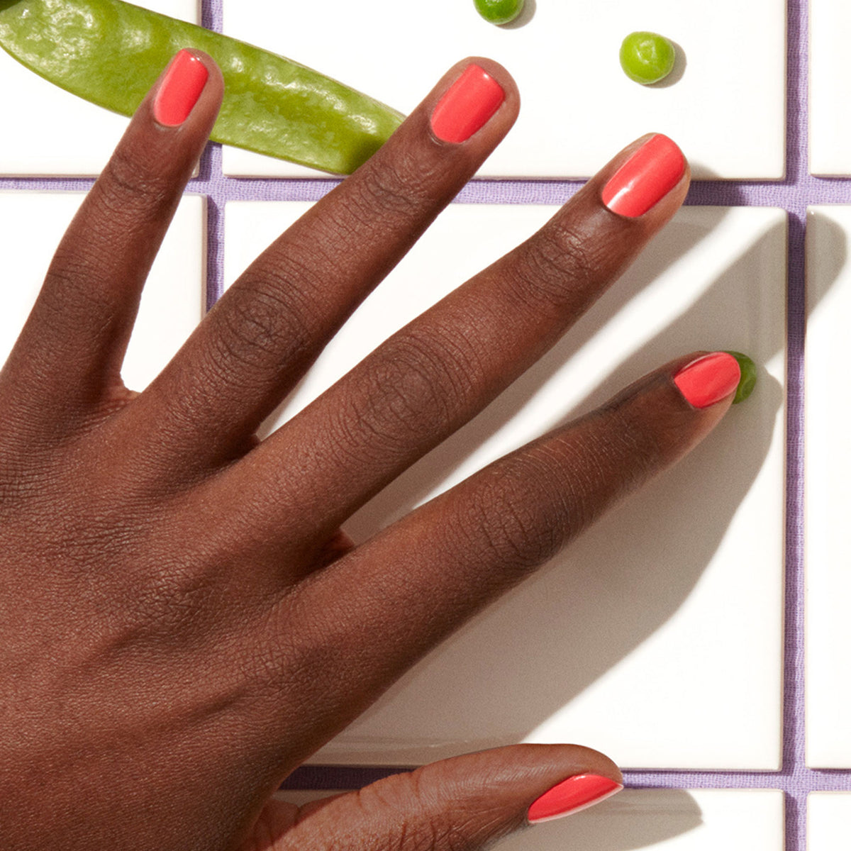 A hand with dark skin and coral pink Pulp nail polish by Manucurist rests on white tiles, surrounded by green snap peas and green peas from the Green range.