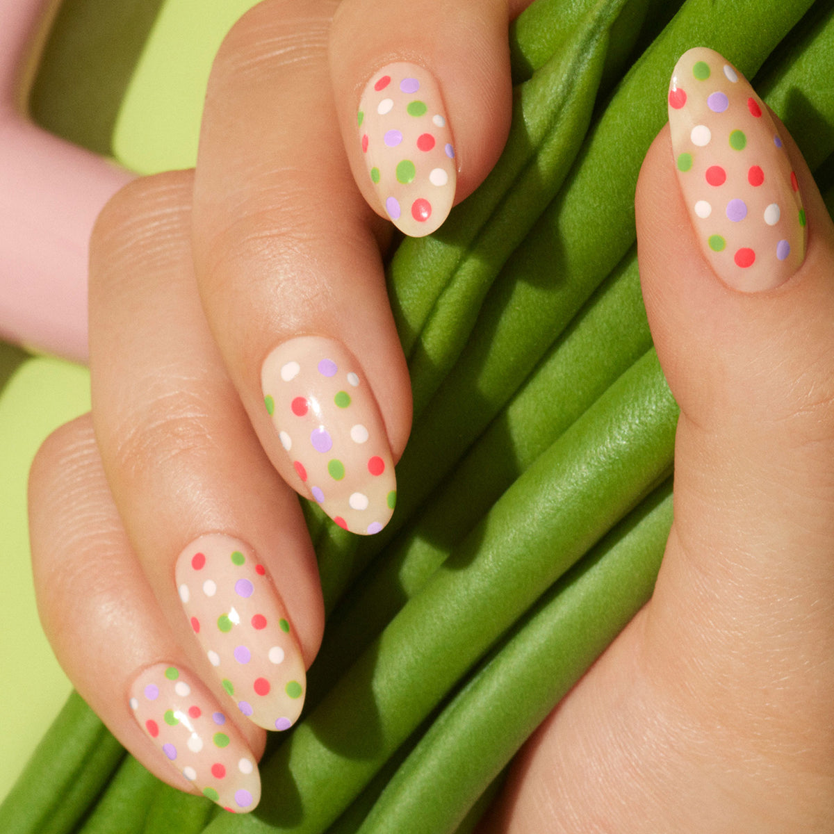Une main aux ongles en amande peints en nude et décorés de pois verts, roses, blancs et violets à l’aide de Dot it better! de Manucurist repose sur un tissu vert texturé.