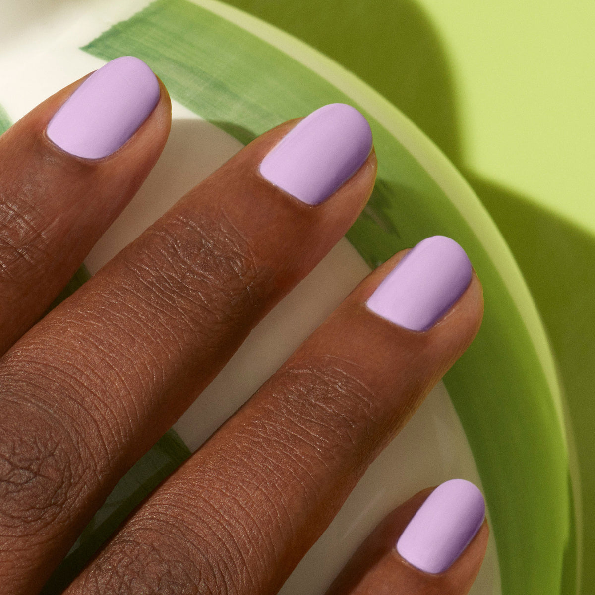 Une main à la peau brun moyen présente des ongles soigneusement manucurés peints avec Lisa Lilas de Manucurist, un vernis gel lilas, sur un fond vert clair et blanc évoquant subtilement les années 90.