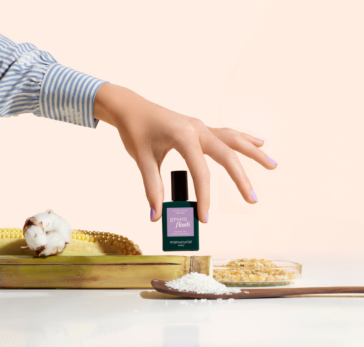 Une main manucurée d'un vernis violet clair tient un flacon étiqueté Green Flash au-dessus d'un épi de maïs, de coton, de céréales et d'une cuillère en bois contenant des granulés blancs, le tout disposé sur une surface claire.