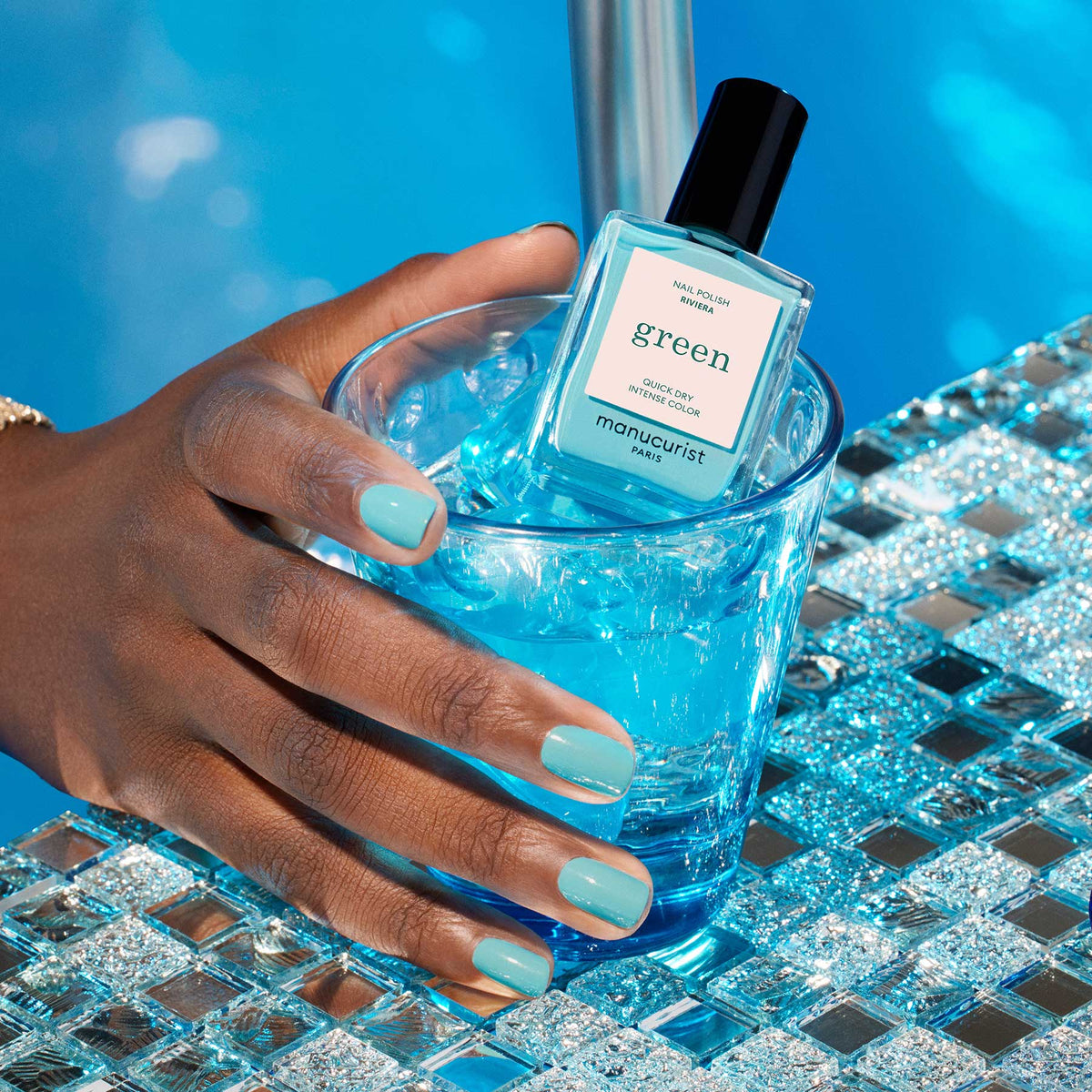 A hand with turquoise blue Riviera by Manucurist nail polish holds a glass of blue iced drink, with the matching nail polish bottle on the ice, set beside a sparkling pool.