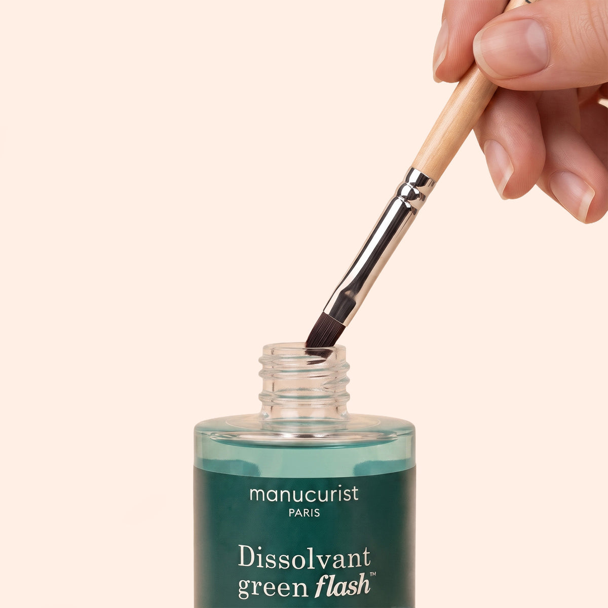 A hand dips a brush into an open bottle labeled manucurist Paris Dissolvant green flash against a pale background.
