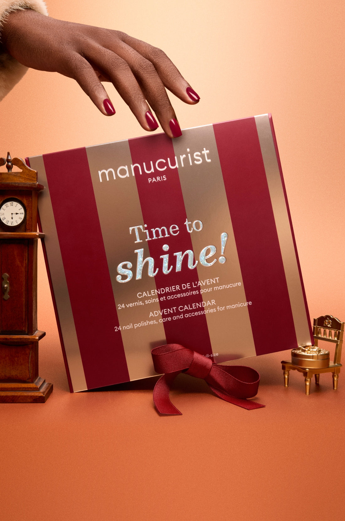 A hand with red nail polish holds a red and gold Manucurist advent calendar box reading Time to shine! near a clock, a bow, and a tiny chair, all on a warm peach background.