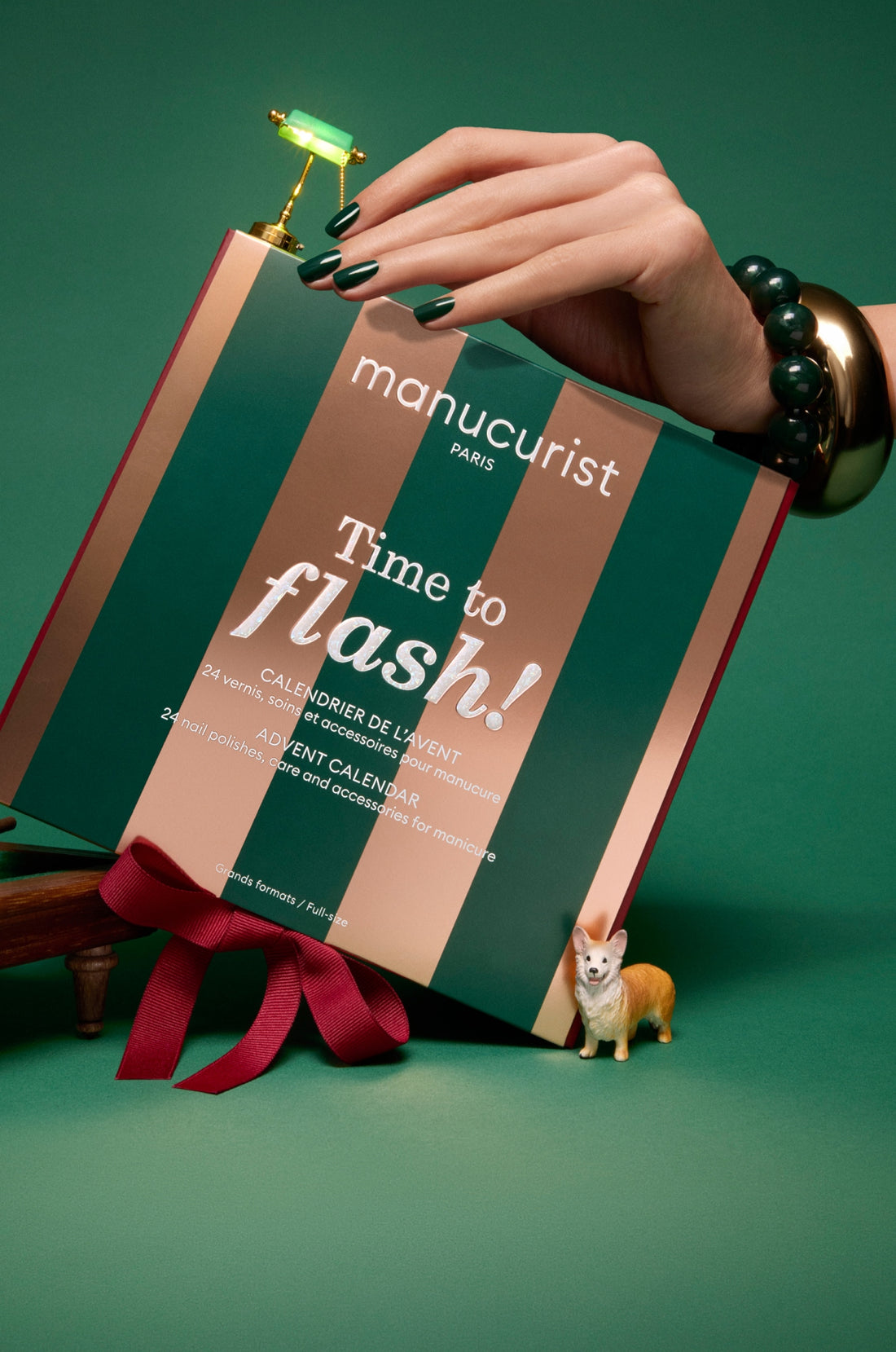 A hand with dark green nails holds a Manucurist Paris Time to flash! advent calendar box, decorated with gold stripes, a red ribbon, and a small toy corgi standing in front on a green surface.