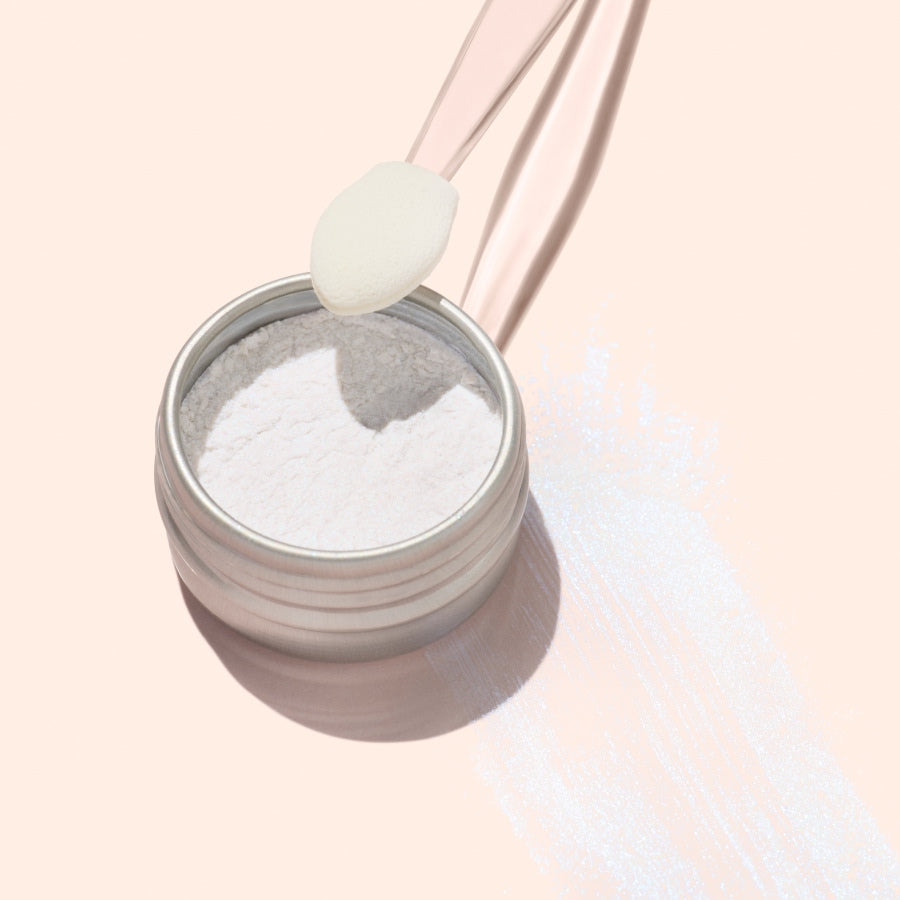 A small round tin of Manucurist Blue Glazed Powder rests on a peach surface. A translucent spatula holds a scoop above the tin, with pearly blue shimmer powder—ideal for adding brilliance to any manicure—scattered nearby.
