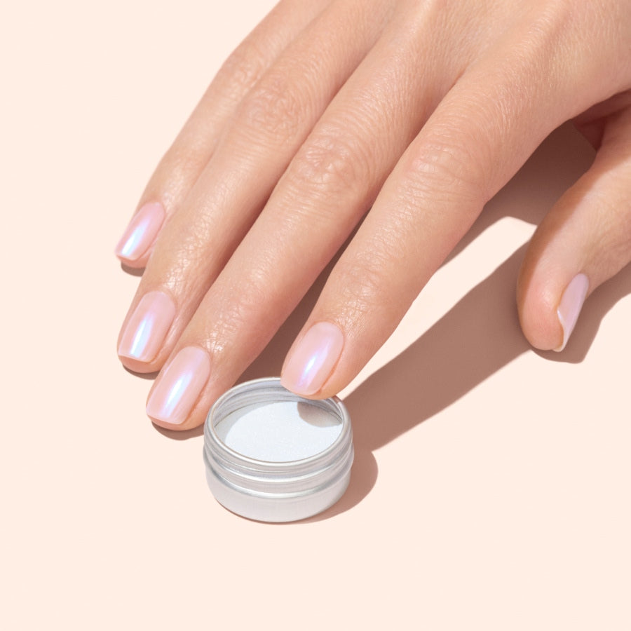 A hand with short, neatly manicured nails painted in a pearly blue shimmer rests beside an open small silver container of Manucurist Blue Glazed Powder on a pale background.