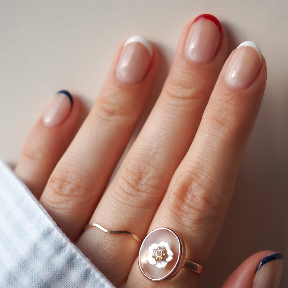 A hand with natural, short nails in a French manicure using Manucurist’s Red, White  Blue Trio and gold flower and thin band rings—perfectly matched with bold red, white, and blue eyeshadow.