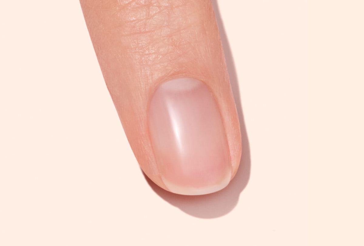 A close-up view of a single fingernail on a finger with natural, healthy, and clean nails against a pale, neutral background.