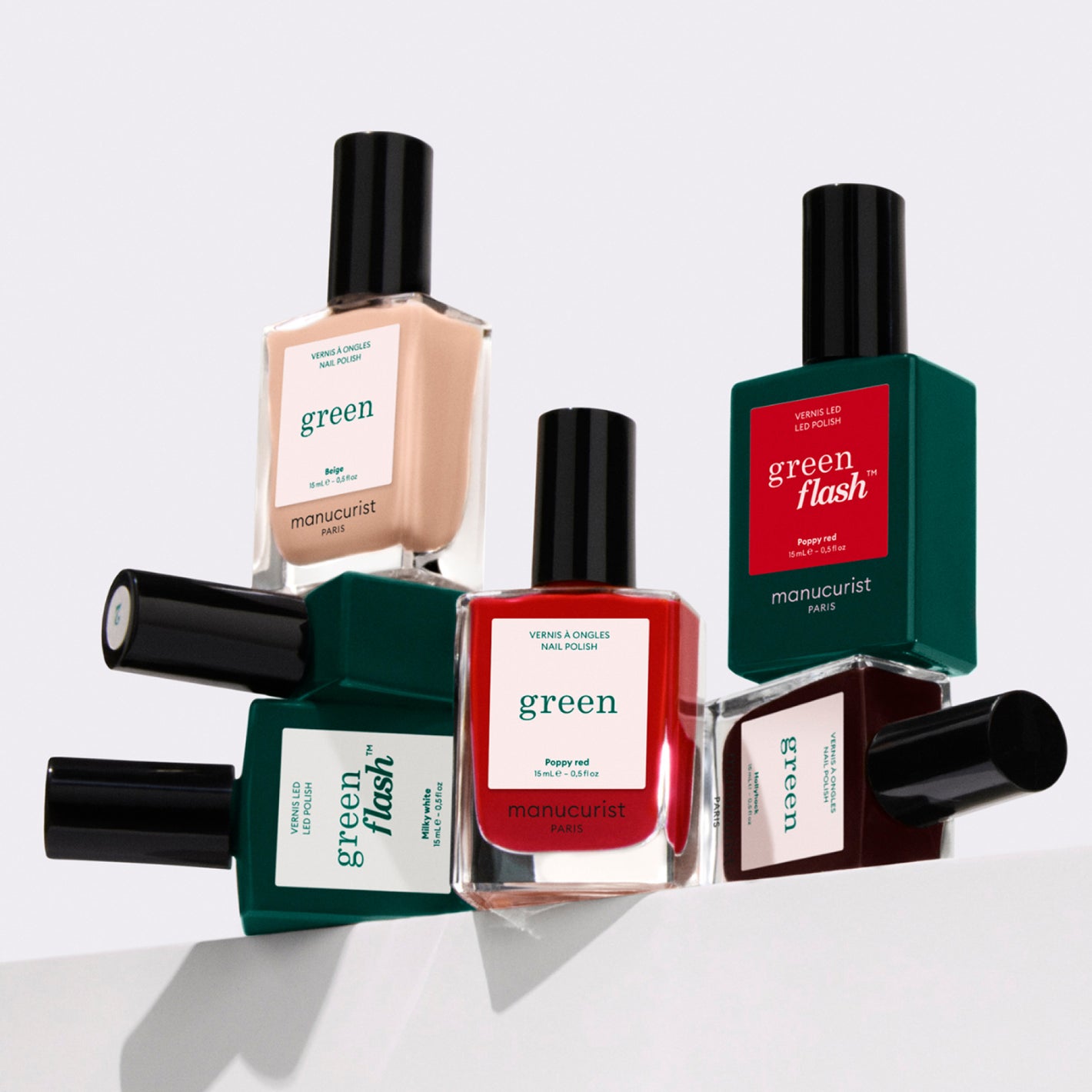 Five bottles of Manucurist nail polish in assorted colors are arranged in a creative, stacked display against a white background. The labels read green and green flash.