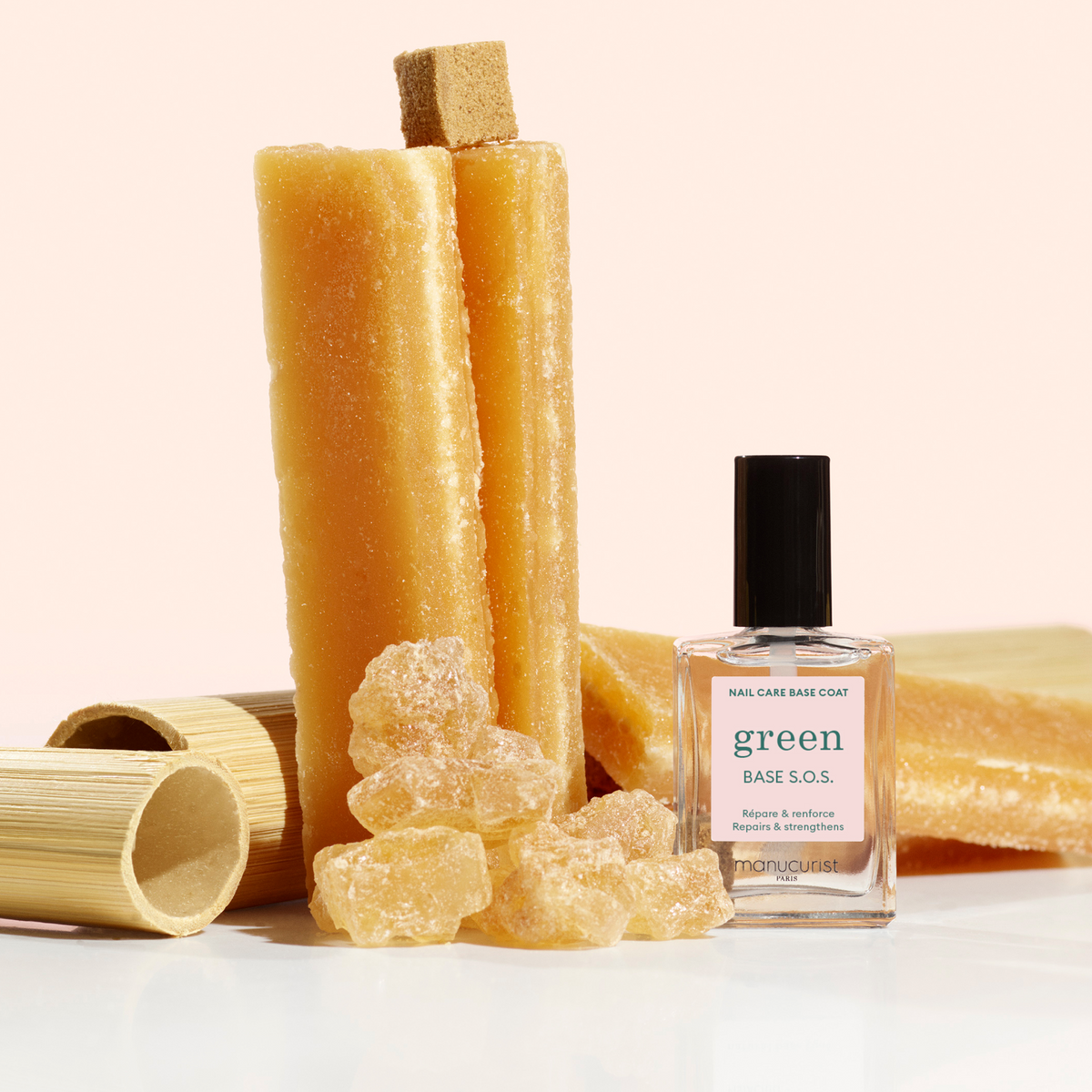 Un flacon de soin pour ongles Manucurist Green Base S.O.S. est posé devant des bâtonnets de sucre ambré, des cristaux et des tubes de bambou sur un fond blanc.